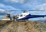 Pilot standing next to helicopter on crest of hill beside a bulldozer.