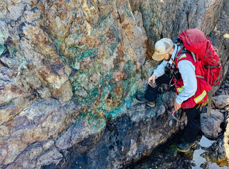 A geologist collects a sample from a green-stained rock face.