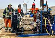 Drillers stand behind a large engine that powers a mineral exploration drill.