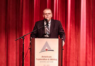 Mining executive behind a podium on a stage backed by red velvet curtains.