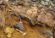 Three rock hammers lie on an orange-stained mineralized outcrop at MAN.