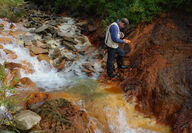 Geologist samples an outcrop of brick red rock along a stream in Alaska.