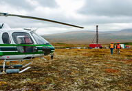 Workers in high visibility vests and hardhats walk from a helicopter to a drill.